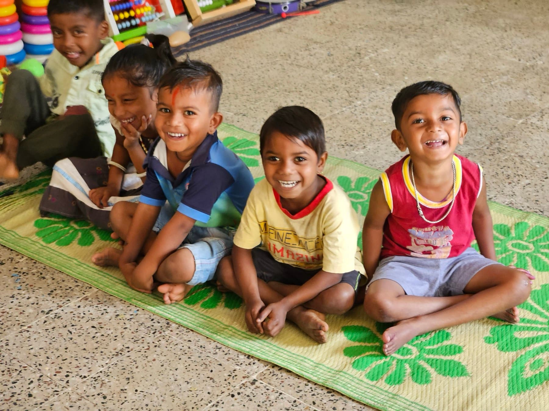 Five young children sit together on a mat, laughing, during a session at their anganwadi.