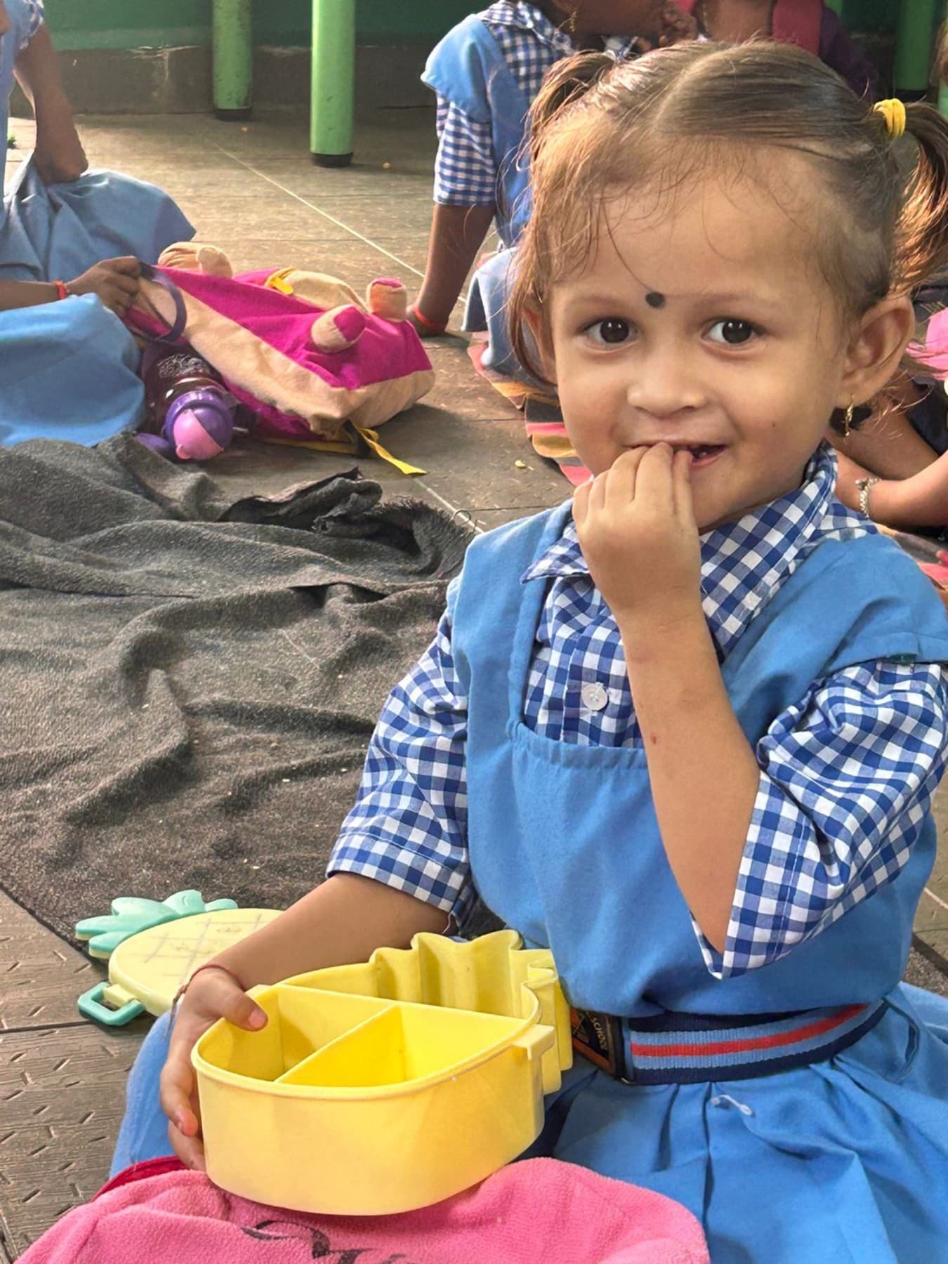A small girl in her checked pinafore holds her lunch box and smiles shyly.