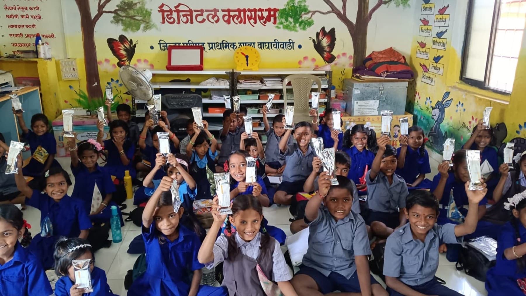 A full classroom of children raise their nutrition sachets together.